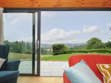 A living area with a blue armchair and a red sofa with cushions facing a sliding glass door opening to a patio and green landscape at Sandsgate Lodge in Spinster's Rock near Chagford
