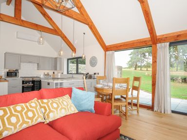 An open plan living area with a red sofa, wooden dining table and chairs, gray kitchen cabinets, and large windows showing green outdoors at Sandsgate Lodge Spinster's Rock near Chagford