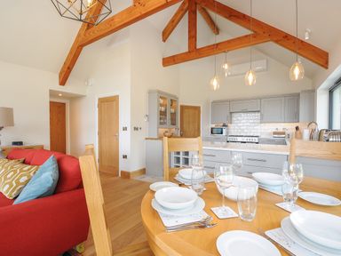 A dining area with a wooden table set with white dishes and glassware next to a kitchen with grey cabinets and wooden ceiling beams at Sandsgate Lodge Spinster's Rock near Chagford