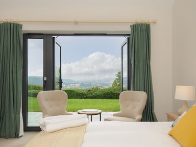 A bedroom with bed lamps two chairs a small table and open doors to a garden and countryside view at Sandsgate Lodge Spinster's Rock near Chagford