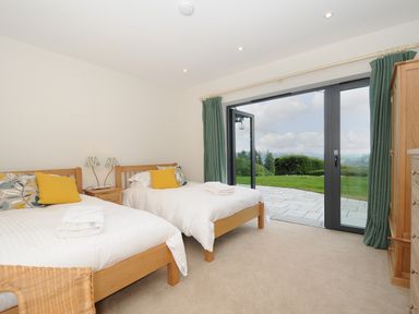 A bedroom with two single beds with yellow cushions and a view of a garden through glass doors at Sandsgate Lodge Spinster's Rock near Chagford