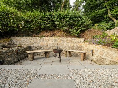 An outdoor seating area with wooden benches and a fire pit surrounded by stone walls at Sandsgate Lodge Spinster's Rock near Chagford
