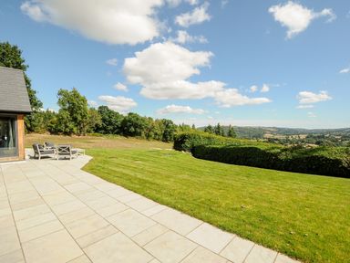 A patio with chairs and a table next to a lawn with trees and countryside in the background at Sandsgate Lodge in Spinster's Rock near Chagford