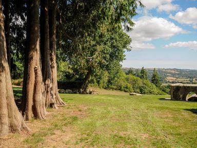 A grassy outdoor area with several large trees on the left some rocks and a stone arch structure on the right and fields in the distance at Sandsgate Lodge in Spinster's Rock near Chagford