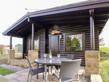 An outdoor patio with a round glass table and four wicker chairs in front of a wooden cabin with stone pillars at Alwinton Lodge in Longframlington