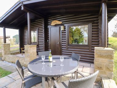 A patio with a round glass table, four chairs, a bottle of wine, and two wine glasses outside a log cabin at Alwinton Lodge in Longframlington