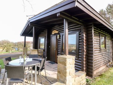 A log cabin with a covered porch and outdoor table with chairs outside at Alwinton Lodge in Longframlington