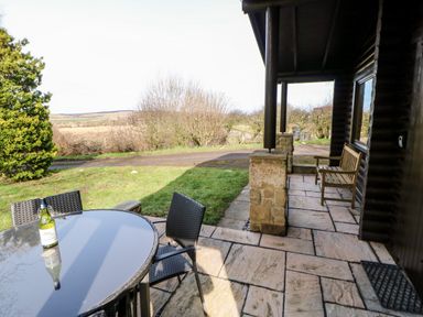 An outdoor patio with a glass table and chairs a bottle on the table a wooden bench on a covered porch at Alwinton Lodge in Longframlington