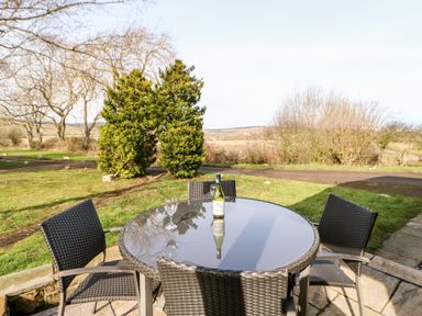 An outdoor patio with a round glass table, four wicker chairs, a bottle of wine, and a wine glass at Alwinton Lodge in Longframlington