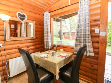 A dining table with two black chairs and place settings next to a window with checkered curtains in a wooden cabin at Alwinton Lodge in Longframlington