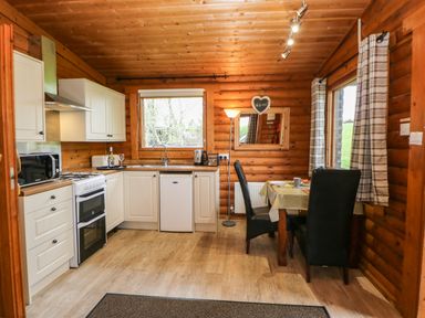 A kitchen and dining area with white cabinets a stove a fridge a table with two chairs and wooden walls at Alwinton Lodge in Longframlington
