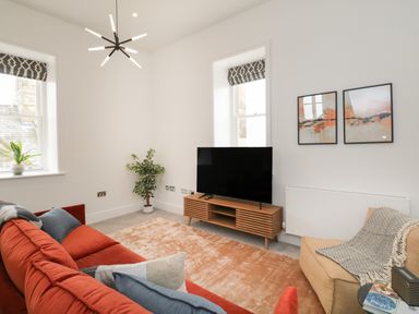 A living room with a red sofa blue and gray cushions a beige armchair a wooden TV stand a flat screen TV two abstract paintings a plant and two windows at William Tower Suite Stone Cross Mansion in Ulverston