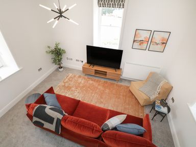 A living room with a red sofa, a tan armchair, a TV on a wooden stand, a plant, and two paintings on the wall at William Tower Suite Stone Cross Mansion Ulverston