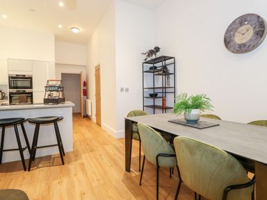 A dining area with green chairs around a table and a kitchen with stools at William Tower Suite Stone Cross Mansion in Ulverston