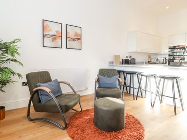 A living area with two chairs, a round ottoman on a round rug, a kitchen counter with four stools, and two paintings on the wall at William Tower Suite Stone Cross Mansion in Ulverston