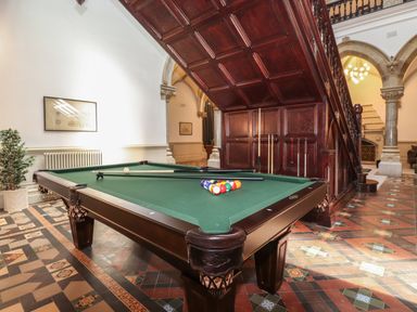 A billiard table with cues under a large wooden staircase in a tiled room at William Tower Suite Stone Cross Mansion in Ulverston
