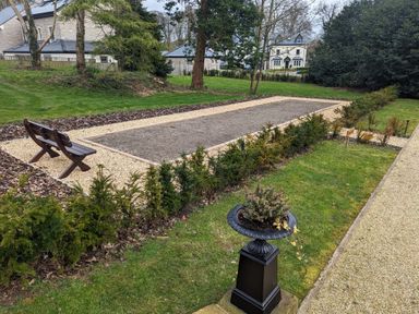 An outdoor gravel bocce ball court with wooden benches surrounded by trees and plants at William Tower Suite Stone Cross Mansion Ulverston
