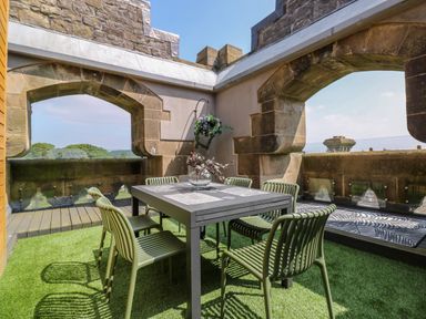 An outdoor dining area with a table and six chairs on artificial grass surrounded by stone archways at William Tower Suite Stone Cross Mansion in Ulverston