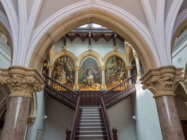 A grand staircase with dark wood banisters and a carpet runner under an arched ceiling with three large paintings at The Elizabeth Suite Stone Cross Mansion in Ulverston