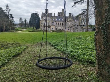 A black circular swing hanging from a tree with a large stone mansion and gardens in the background at The Elizabeth Suite Stone Cross Mansion in Ulverston