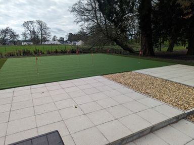 An outdoor putting green with golf flags surrounded by tiled and gravel areas with trees and houses in the background at The Elizabeth Suite Stone Cross Mansion in Ulverston