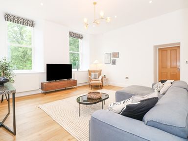 A living room with a gray sectional sofa a black coffee table wooden TV stand and two large windows at The Elizabeth Suite Stone Cross Mansion in Ulverston