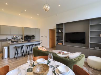 An open plan living area with a kitchen island bar with stools a dining table with place settings a green sofa and a wall-mounted television at The Alfred Suite Stone Cross Mansion in Ulverston