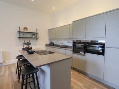A kitchen with an island, three black stools, built-in ovens, cabinets, and wall shelves at The Alfred Suite Stone Cross Mansion in Ulverston