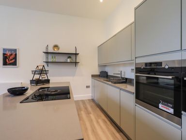 A modern kitchen with grey cabinets a built in oven an induction cooktop a countertop with a bowl and a tiered basket a wall shelf with decor items and a framed picture on the wall at The Alfred Suite Stone Cross Mansion Ulverston