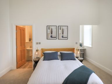 A bedroom with a bed a yellow headboard and blue pillows a door leading to a bathroom and a window with a small desk at The Alfred Suite Stone Cross Mansion Ulverston