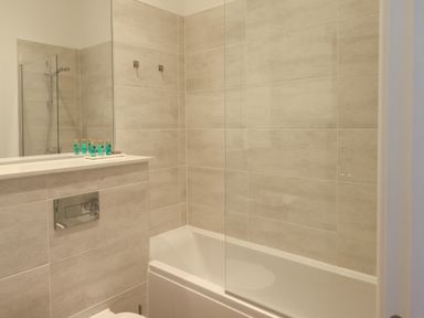A bathroom with a bathtub shower combination a glass shower screen and toiletries on a shelf at The Alfred Suite Stone Cross Mansion in Ulverston