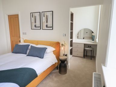 A bedroom with a bed with blue pillows and a yellow headboard a vanity with a round mirror and a stool at The Alfred Suite Stone Cross Mansion in Ulverston