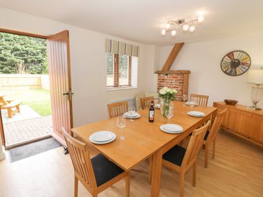 A dining room with a wooden table set with plates glasses a bottle and flowers and an open door leading outside at The Wheat House in Welford-On-Avon