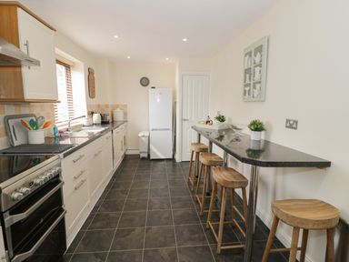 A kitchen with white cabinets black countertops stools along a bar and a white refrigerator at The Wheat House in Welford-On-Avon