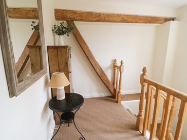 A stairway landing with wooden railing a black round table with a lamp a wooden cabinet a large wall mirror and exposed wooden beams at The Wheat House in Welford-On-Avon