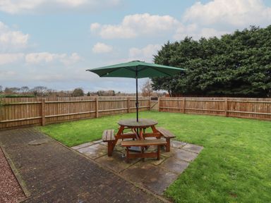 A garden with a wooden picnic table and green umbrella on a paved area surrounded by grass and a wooden fence at The Wheat House in Welford-On-Avon