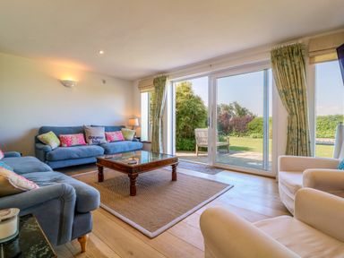 A living room with blue sofas white armchairs a wooden coffee table and glass doors leading to a garden at Alpha in St Minver