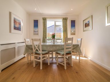 A dining room with a table covered by a cloth and six chairs around it with curtains on a window and framed pictures on the walls at Alpha in St Minver