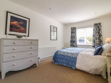A bedroom with a bed covered in blue and white bedding a gray dresser with a framed picture on the wall and a window with patterned curtains at Alpha in St Minver