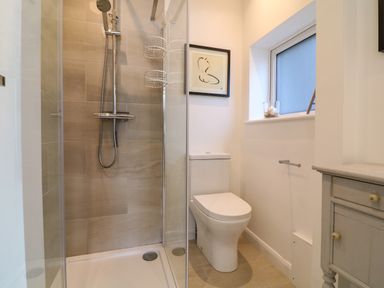 A bathroom with a shower, toilet, window, framed artwork, and a small cabinet at Alpha in St Minver