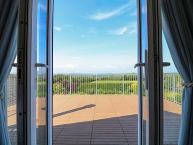 An open glass door leading to a wooden deck with metal railing overlooking green fields and a blue sky at Alpha in St Minver