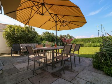 An outdoor patio with a large table and chairs under a yellow umbrella with garden and lawn at Alpha in St Minver