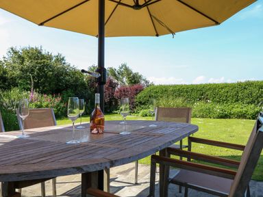 A wooden outdoor table with wine glasses and a bottle under a large umbrella in a garden at Alpha in St Minver