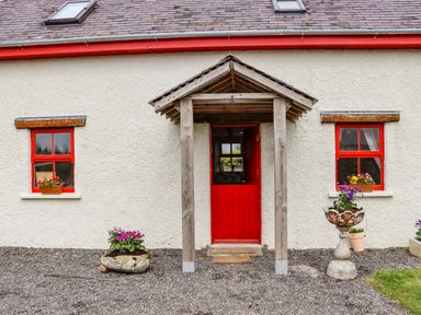 The exterior front of a house with a red door and two windows with red frames and flower pots at Cob Cottage in Kilmore County Wexford
