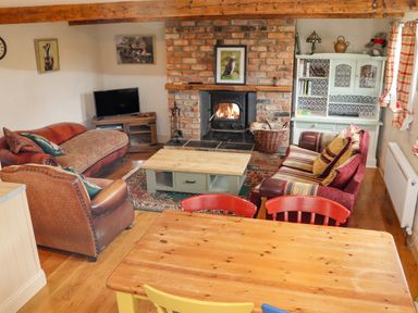 A living room with a brick fireplace a wood burning stove sofas a wooden coffee table a dining table with colorful chairs and a white cabinet at Cob Cottage in Kilmore County Wexford