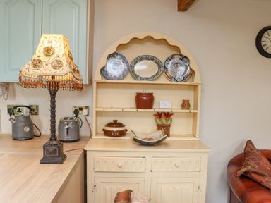 A corner of a kitchen with a cream wooden cabinet displaying decorative plates and pottery next to a countertop with a lamp kettle and toaster at Cob Cottage in Kilmore County Wexford