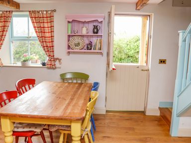 A dining area with a wooden table and colorful chairs next to a window with checkered curtains and a half-open door at Cob Cottage in Kilmore County Wexford