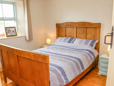 A bedroom with a wooden bed frame striped bedding two bedside tables with lamps and a window with a painting on the sill at Cob Cottage in Kilmore County Wexford