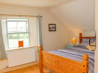 A bedroom with a wooden bed a bedside table with a lamp a window with light curtains and a small chest on the windowsill at Cob Cottage Kilmore County Wexford