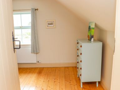 A small room with a wooden floor a window with curtain a radiator a chest of drawers and a painting on the chest at Cob Cottage in Kilmore County Wexford